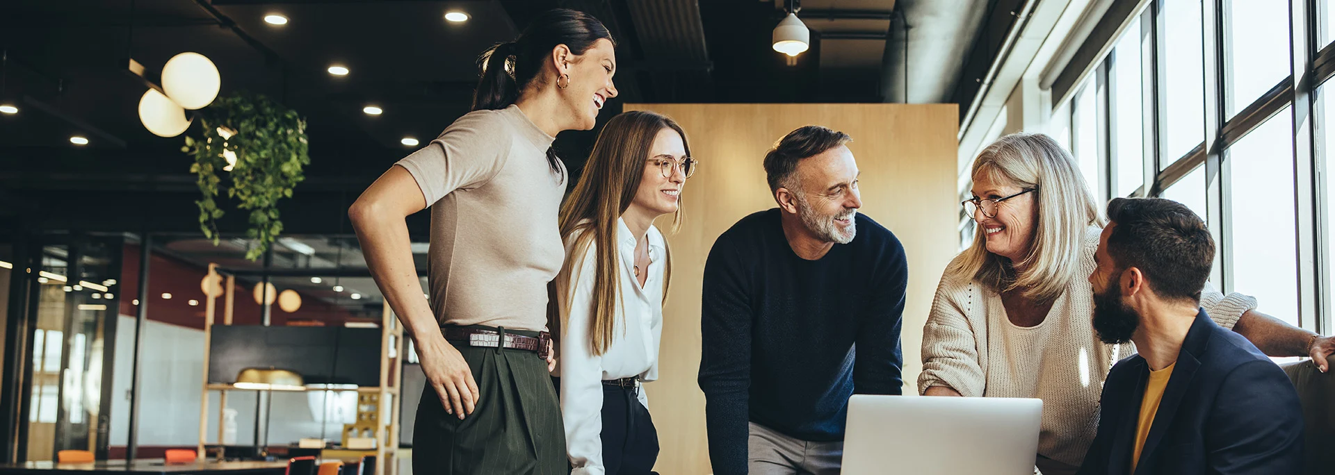 Eine Gruppe von fünf Geschäftsleuten unterschiedlichen Alters lacht und diskutiert gemeinsam in einem modernen Büro vor einem Laptop – forever young Wiederverkäufer-Programm